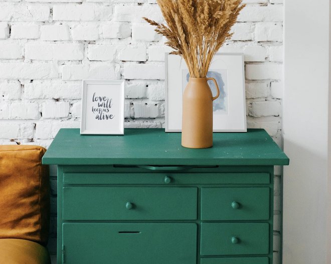 Organized furniture, a green cabinet, two white picture frames, and a beige vase holding wheat